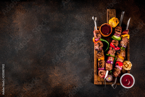 Fresh, home-cooked on the grill fire meat beef shish kebab with vegetables and spices, with barbecue sauce and ketchup, on a dark background on a wooden cutting board above copy space