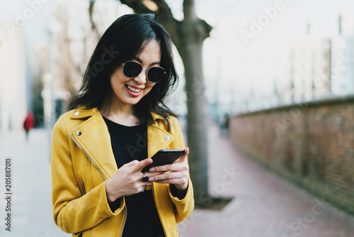 Half length portrait of a beautiful smiling asian hipster girl in a sunglasses with long dark hair wearing yellow leather jacket surfing web on a mobile phone while standing on a blurred background.