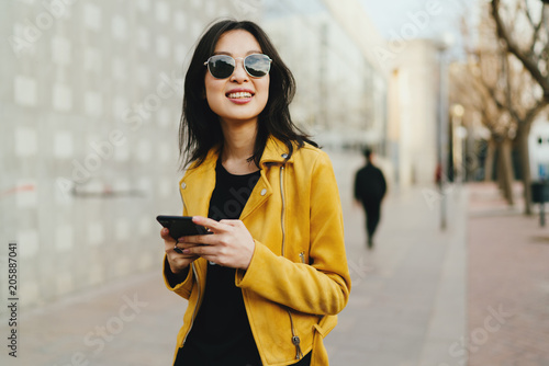 Half length portrait of a beautiful smiling asian woman in a sunglasses with long dark hair wearing yellow leather jacket chatting online by a mobile phone while standing on blurred street background.