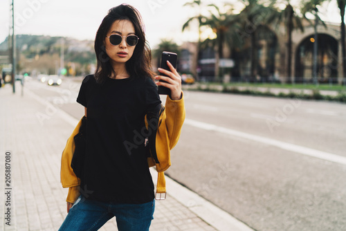 Stylish asian woman with long dark hair in sunglasses wearing blank black t-shirt and yellow leather jacket taking selfie by a mobile phone while standing on a blurred urban background.