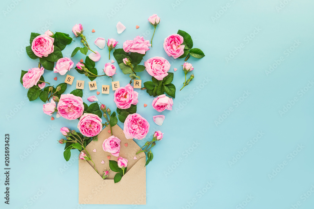 Close up reative layout with pink tea rose flowers, green leaves, candy hearts fly out of craft paper envelope on the light blue background. Word Summer lettering by wooden letters. Text space.