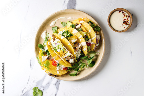 Variety of vegetarian corn tacos with vegetables, green salad, chili pepper served on ceramic plate with cream sauce over white marble background. Top view, copy space.