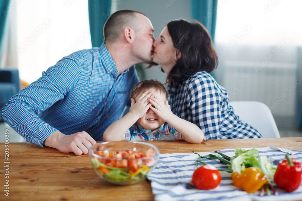 Young husband and wife kiss, son smiling, sitting at the table, family. Concept of a happy family healthy eating Young husband and wife kiss, son smiling, sitting at the table, family. Concept of a happy family healthy eating