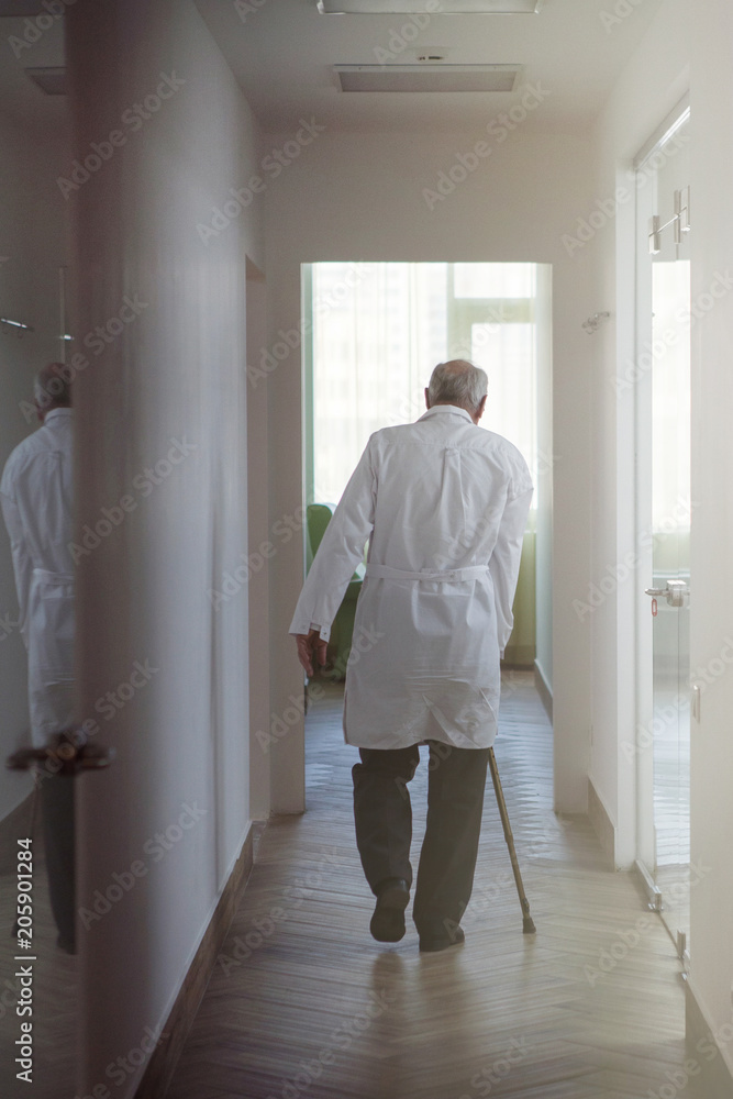 An elderly doctor in a white coat with a cane leaves into the distance along the corridor
