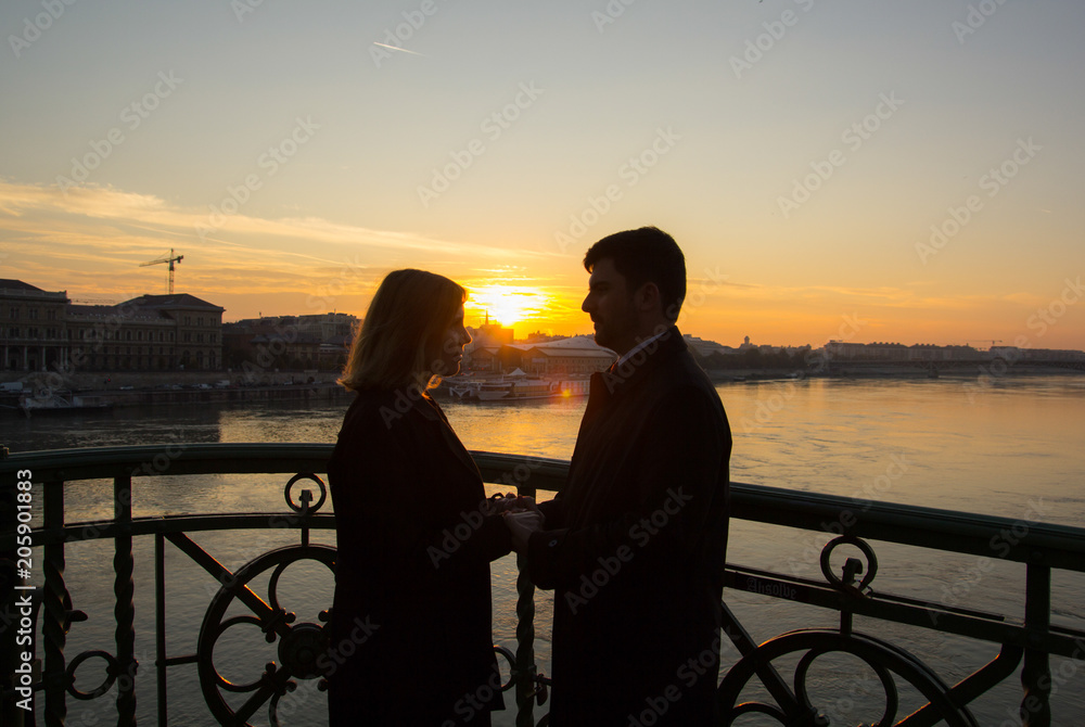 Couple in love is standing on the liberty bridge over the river Danube in Budapest. Sunrise in the big city. Dark silhouettes of man and woman holding hands in urban landscape. Sun kiss.