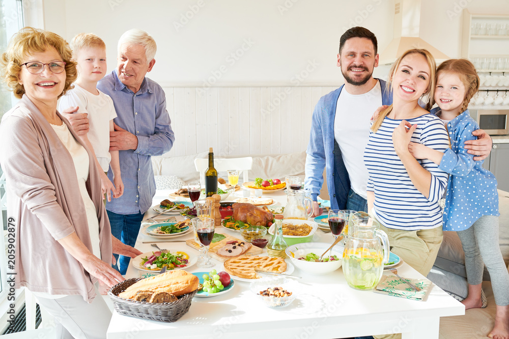 Portrait of happy two generation family enjoying dinner posing round festive table with delicious dishes and smiling at camera during holiday celebration in modern sunlit apartment, copy space