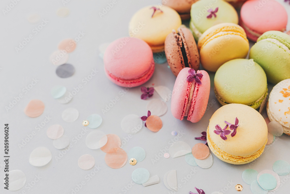 tasty pink, yellow, green and brown macaroons on trendy pastel gray paper with lilac flowers and confetti. space for text. delicious colorful macaroons candy for party. yummy background tasty pink, yellow, green and brown macaroons on trendy pastel gray paper with lilac flowers and confetti. space for text. delicious colorful macaroons candy for party. yummy background