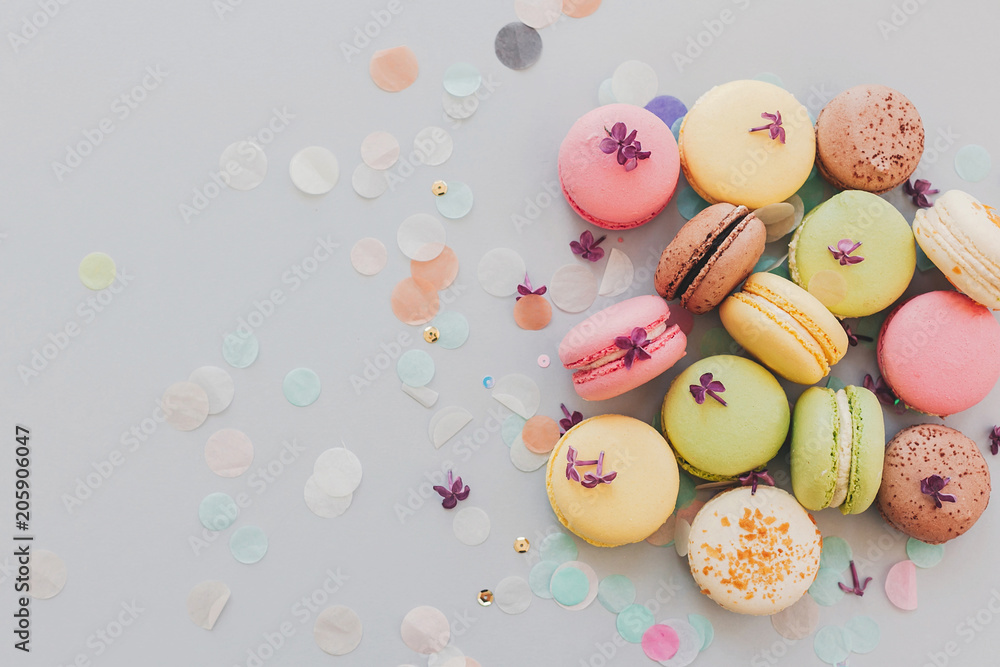 delicious colorful macaroons on trendy pastel gray paper with lilac flowers and confetti, flat lay. tasty pink, yellow, green and brown macaroons. space for text. candy for party