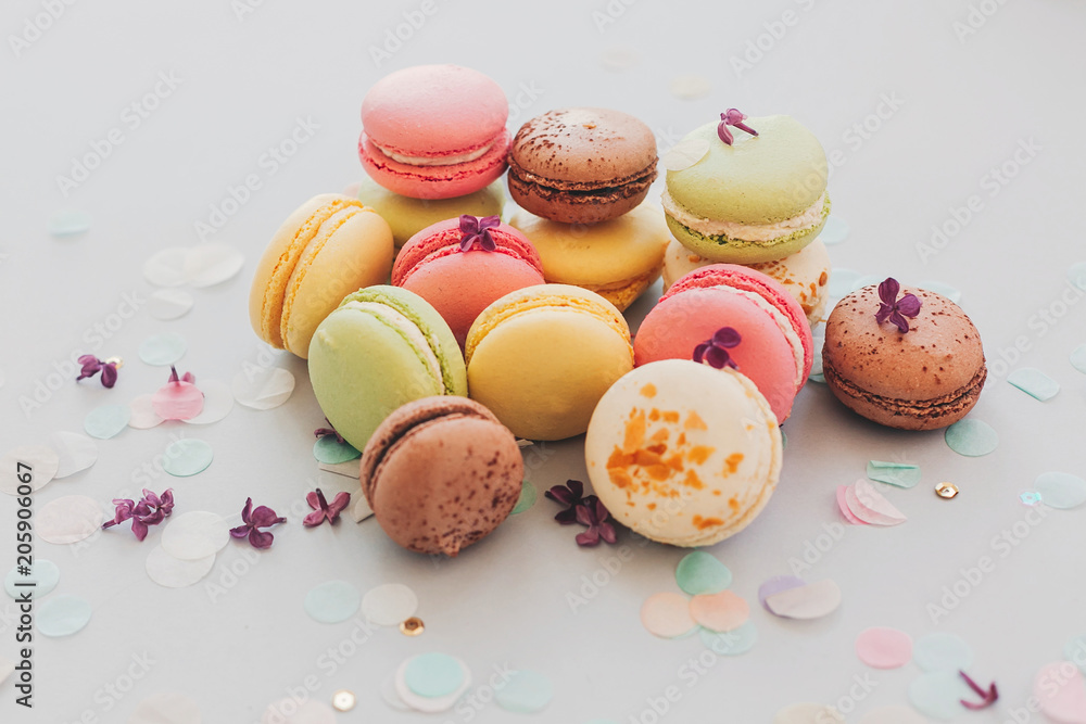 tasty pink, yellow, green and brown macaroons on trendy pastel gray paper with lilac flowers and confetti. space for text. delicious colorful macaroons candy for party. yummy background tasty pink, yellow, green and brown macaroons on trendy pastel gray paper with lilac flowers and confetti. space for text. delicious colorful macaroons candy for party. yummy background