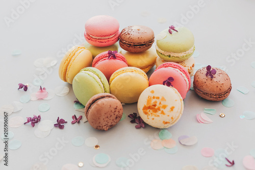 tasty pink, yellow, green and brown macaroons on trendy pastel gray paper with lilac flowers and confetti. space for text. delicious colorful macaroons candy for party. yummy background