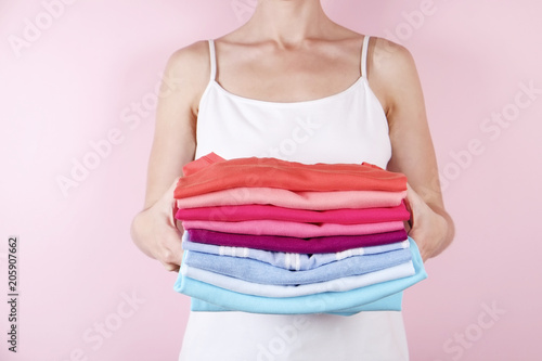 Close up of attractive young woman holding stack of perfectly folded multicolor shirts. Female w/ pile of different color clothing in her hands on pale pink background. Laundry concept. Copy space.