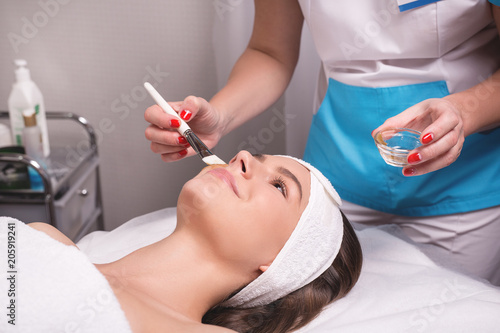 Stunning young woman with hair fixed behind,clean fresh skin naked shoulders wearing white bath robe and hair wrap, doing cosmetic procedure, close up, microdermabrasion