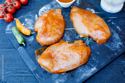 Raw marinated chicken breast fillets on  stone plate tray with BBQ sauce in white dish on dark old wooden rustic background, prepatation for grilling or smoking, top view, overhead photo