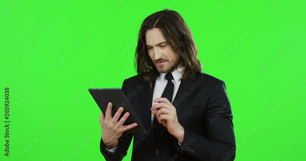 Portrait shot of the young Caucasian businessman in the black suit and tie scrolling and taping on the tablet, then doing yes gesture on the chroma key background. Green screen. Portrait shot of the young Caucasian businessman in the black suit and tie scrolling and taping on the tablet, then doing yes gesture on the chroma key background. Green screen.