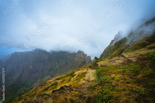 Trekking path beside the mountain peak chain overgrown with verdant grass and cultivated plants. Foggy clouds moving over the Mountain edge. Xo-Xo Valley. Santo Antao Island, Cape Verde Cabo Verde