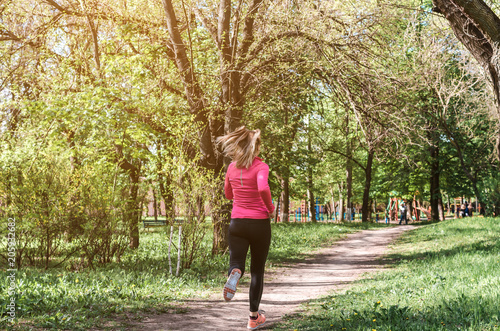 A young athletic girl in a pink and black sports suit jogging along the path in the park in the morning.