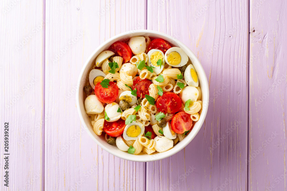 Pasta salad with quail eggs, mozzarella, cherry tomatoes and capers in bowl on purple wooden background Pasta salad with quail eggs, mozzarella, cherry tomatoes and capers in bowl on purple wooden background