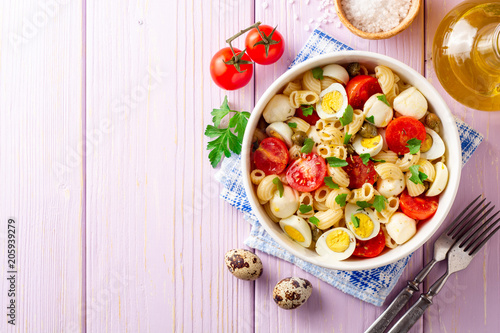 Pasta salad with quail eggs, mozzarella, cherry tomatoes and capers in bowl on purple wooden background