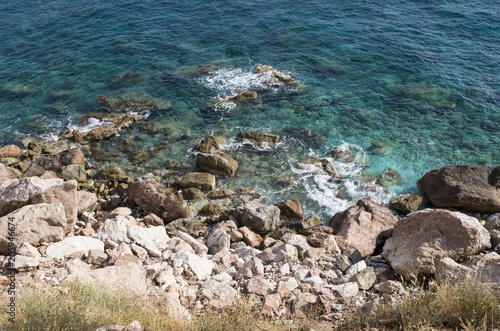 Sea rocky coast with clear water of emerald color and white foam on a sunny day, top view; Blue-green sea water and big rose stones on the seashore. 