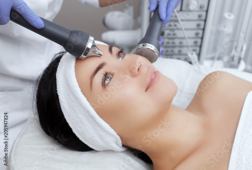 The cosmetologist makes the procedure an ultrasonic cleaning of the facial skin of a beautiful, young woman in a beauty salon.Cosmetology and professional skin care. photo