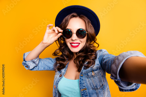 Portrait of trendy pretty tourist with beaming smile shooting self portrait on front camera holding eyelet of glasses on face wearing jeans jacket isolated on yellow background, travel tourism concept