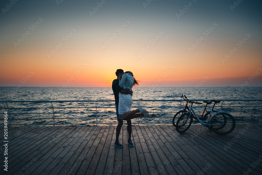 Fototapeta premium Couple of young hipster lovers kissing at the beach at sunrise sky at wooden deck summer time