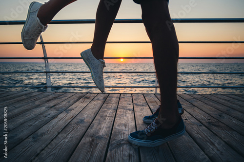 Couple of young hipster lovers kissing, close up on legs and sneakers at the beach at sunrise sky at wooden deck summer time