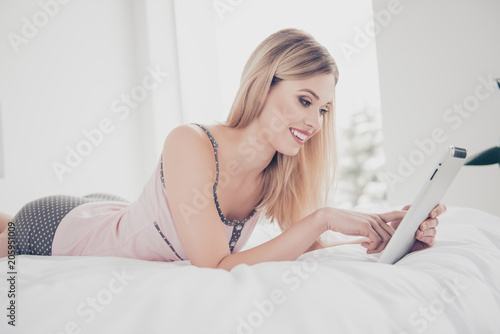 Portrait of attractive, successful, confident, pretty, charming, cheerful, stylish girl lying on bed in pajamas, having tablet in arms, checking email, looking news, photos, using wi-fi internet