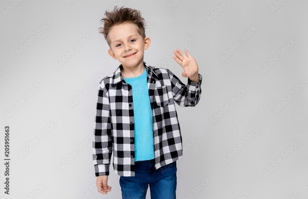 A handsome boy in a plaid shirt, blue shirt and jeans stands on a gray background. The boy smiles and shows the palm A handsome boy in a plaid shirt, blue shirt and jeans stands on a gray background. The boy smiles and shows the palm