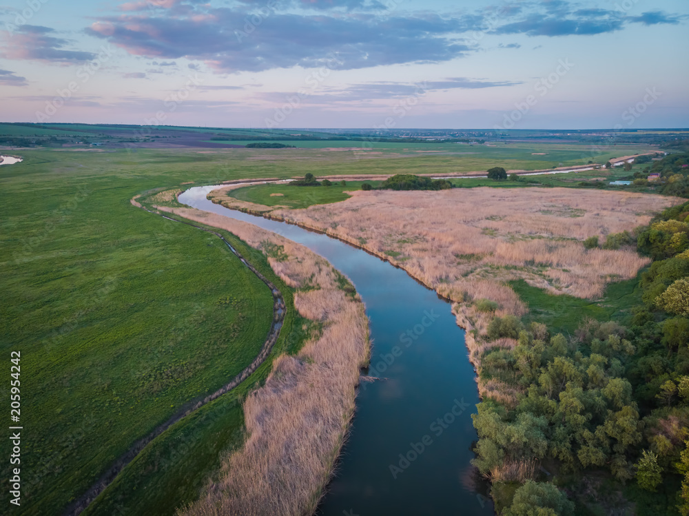 Quiet river flows along the plain. Riversides, overgrown with reeds, green meadows. Beautiful scenery from a bird's eye view at sunset