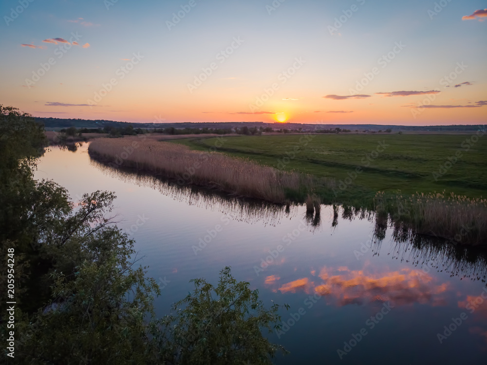 The calm surface of the river and the reflections of clouds, orange sunset, green fields and meadows in a quiet warm summer evening