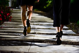 Close up of legs of two young skinny women with heels walking towards the club by night. Black dressing, white way with flowers, crazy party