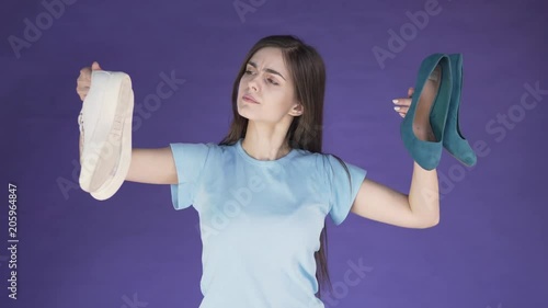 Long-haired attractive girl hesitating between white trainers and green suede high-heeled shoes, isolated shot in purple background, concept of customer's choice