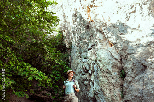 Girl traveler walks through the mountains and woods. He smiles. Green shirt shorts and tan. Straw hat. Photographer is a tourist researcher.