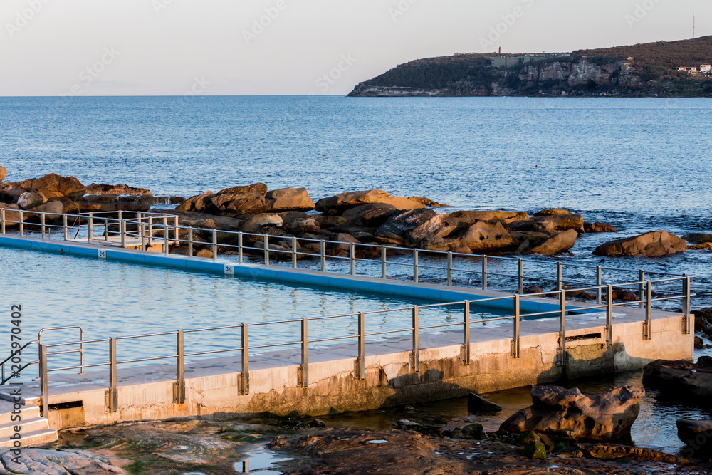 Seaside ocean swimming pool with metal hand rail fencing set against calm blue water and large exposed rocks 