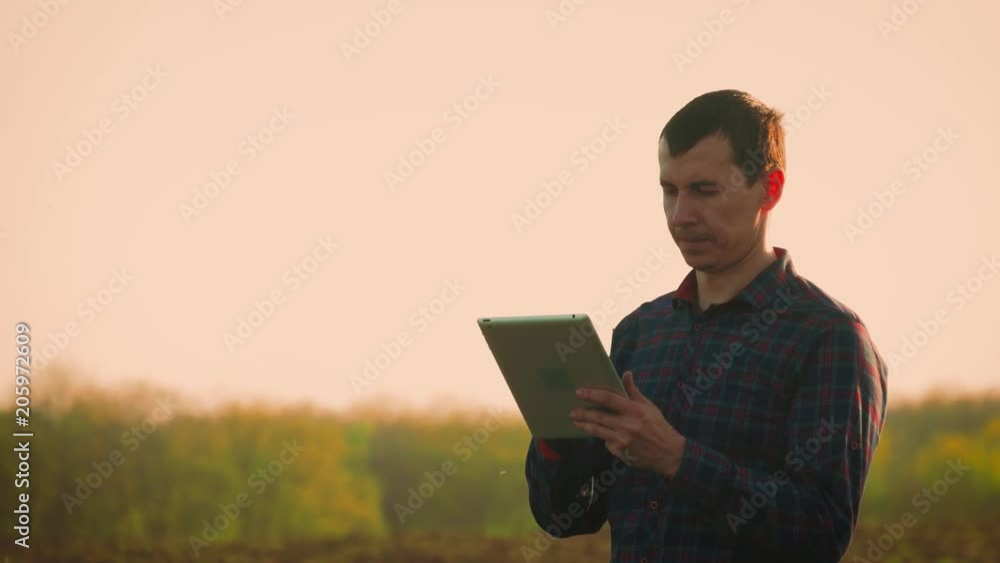Young farmer holding tablet in front of tractor in field. Seasonal agricultural worker. Concept ecology, transport, farmers, clean air, food, bio product. Young farmer holding tablet in front of tractor in field. Seasonal agricultural worker. Concept ecology, transport, farmers, clean air, food, bio product.