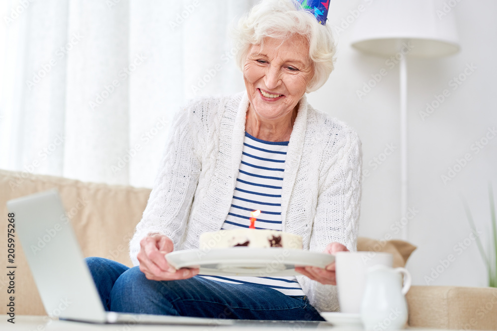 Portrait of happy senior woman showing cake to camera while celebrating birthday with her family via video chat Portrait of happy senior woman showing cake to camera while celebrating birthday with her family via video chat