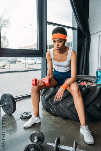 Fototapeta Naklejka Na Ścianę i Meble -  african american sportswoman in headband and wristbands doing exercise with dumbbell while sitting on training tire at gym