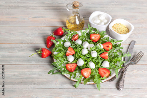Delicious rucola salad with mozzarella, strawberries, olive oil and spices on a wooden background. Healthy foods