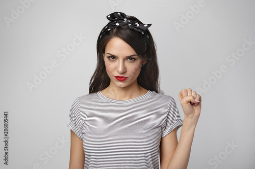 Horizontal shot of attractive young woman feminist wearing bright make up and headscarf having confident look, raising clenched fist as sign of feminism, equal rights, respect and girl power