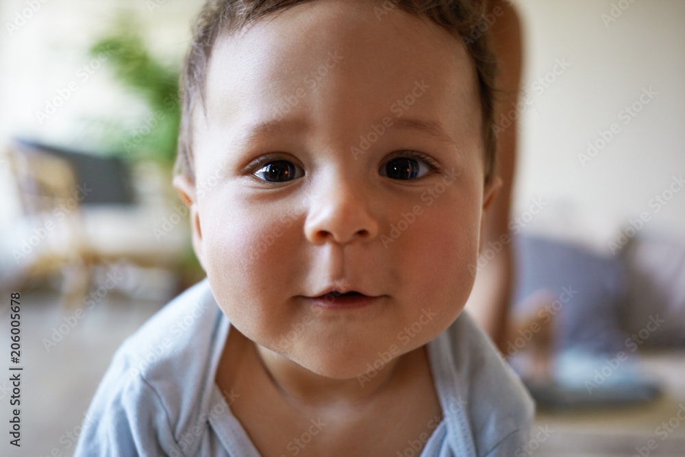Close up highly detailed portrait of adorable chubby infant boy of mixed race appearance having playful expression, looking at camera and smiling while playing at home. Infantry and childhood