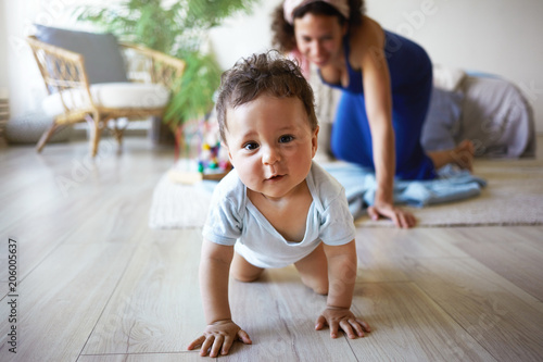 Canvas Print Horizontal shot of cute funny infant crawling along floor from his mother who is standing on her knees in background, trying to catch her baby son, smiling happily