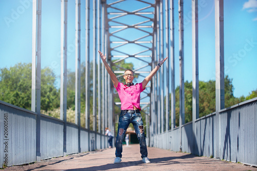Cute emotional man with headphones in glasses in a pink shirt walking on a pedestrian bridge