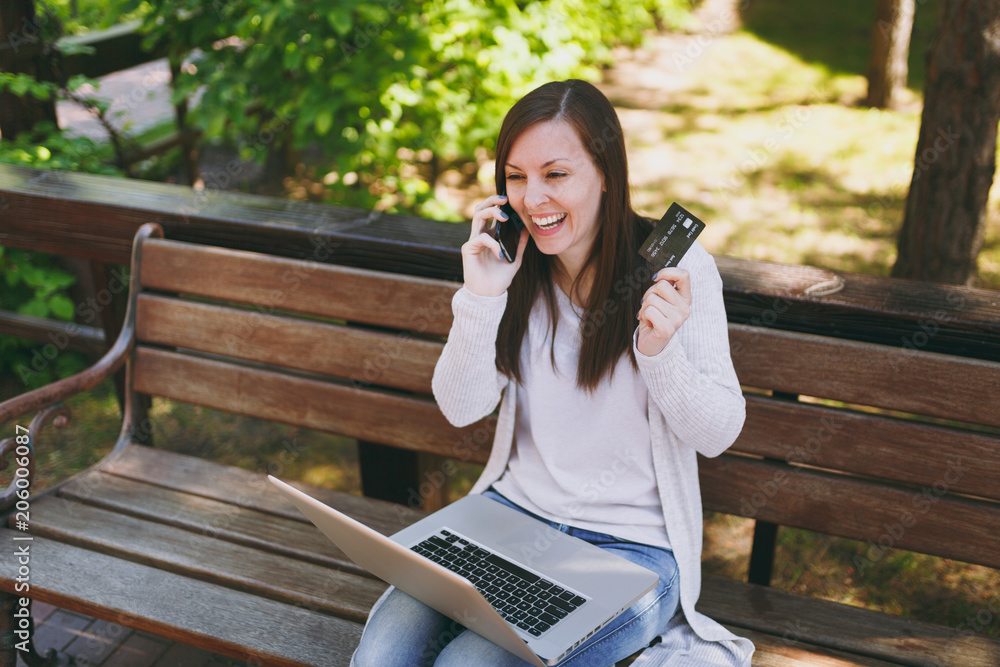 Female holding credit card, talking on mobile phone, having good pleasure conversation. Woman sitting on bench working on modern laptop in street outdoors. Mobile Office. Freelance business concept. Female holding credit card, talking on mobile phone, having good pleasure conversation. Woman sitting on bench working on modern laptop in street outdoors. Mobile Office. Freelance business concept.