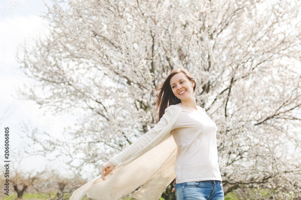 Young happy relaxed smiling beautiful woman in light casual clothes spreading hands standing in city garden or park on blooming tree background. Spring nature, flowers. Lifestyle, leisure concept. Young happy relaxed smiling beautiful woman in light casual clothes spreading hands standing in city garden or park on blooming tree background. Spring nature, flowers. Lifestyle, leisure concept.