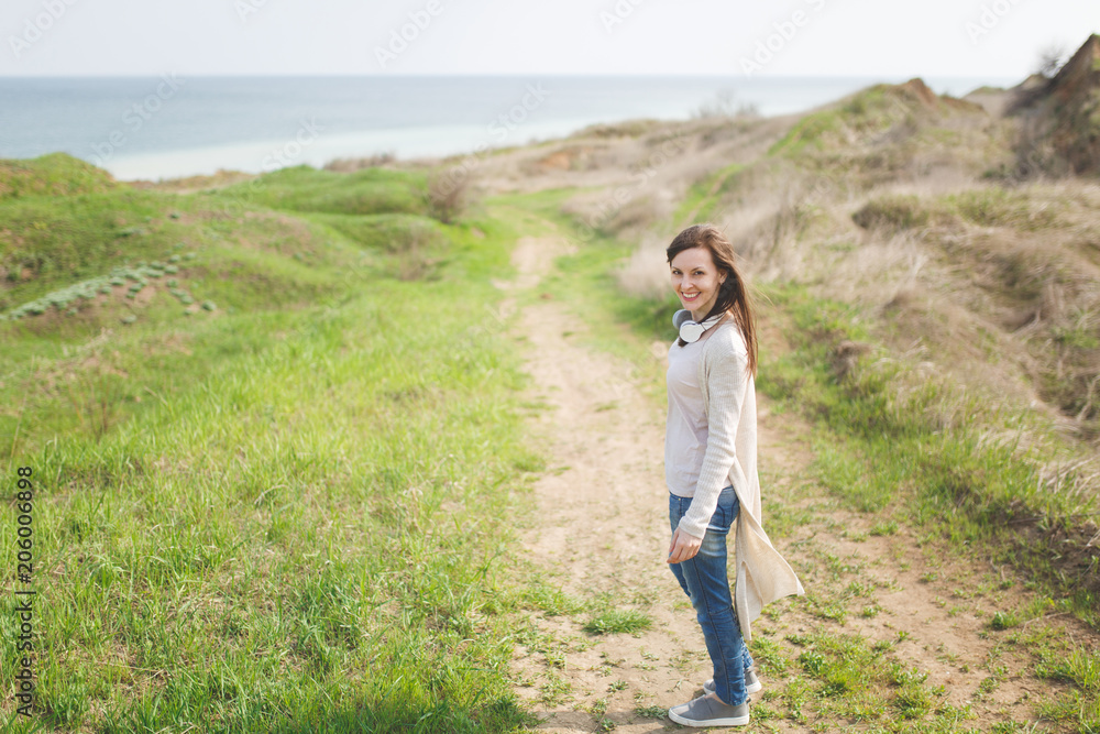 Young brunette smiling beautiful woman in light casual clothes walking along path standing in sunny weather in field near water on green background. Beautiful landscape. Lifestyle, leisure concept.
