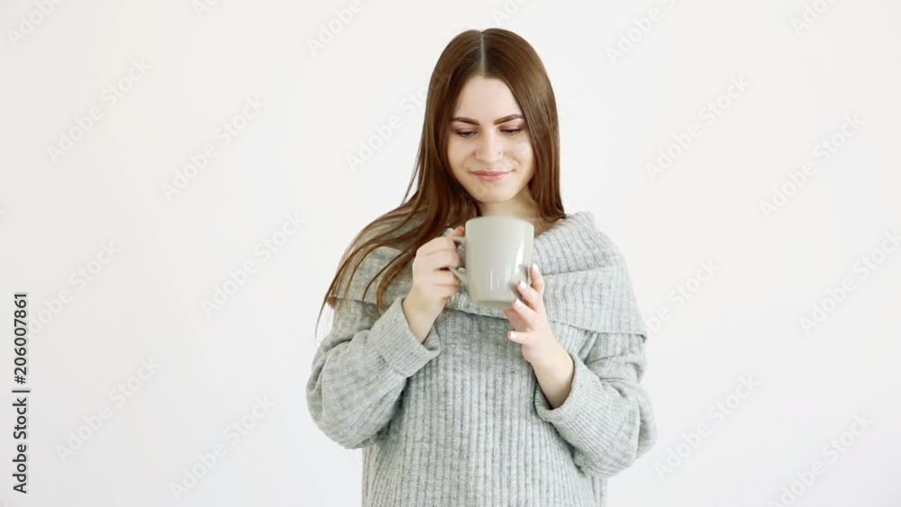 beautiful happy girl of european appearance in a warm sweater drinks hot tea by the white wall. beautiful happy girl of european appearance in a warm sweater drinks hot tea by the white wall.