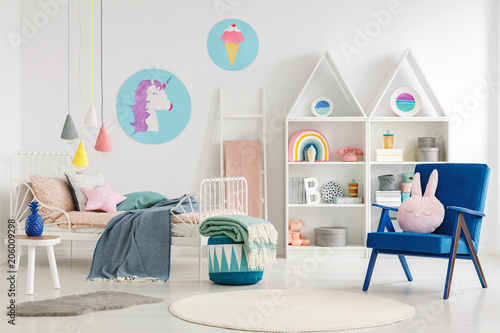 Sweet bedroom interior for a kid with a blue armchair, rabbit pillow, bed, unicorn and ice cream posters and shelves photo
