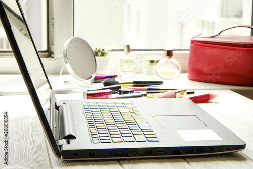 Feminine desktop top with bunch of cosmetics: eye shadow palette, brow mascara, blusher, highlighter and lipstick . Woman's table with blank screen laptop computer and beauty accessories. Background.