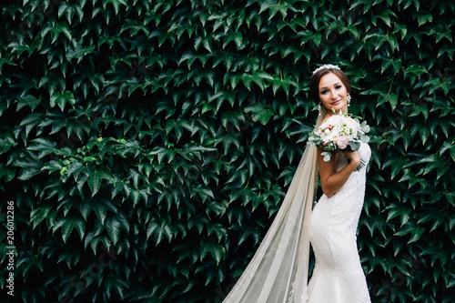 Portrait of a bride in a white dress with a wedding bouquet in the hands, against a background of a green wall of plants photo